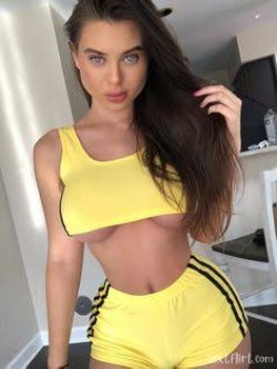 Brunette woman with curvy figure wearing yellow crop top and matching shorts, posing indoors with hand in hair.