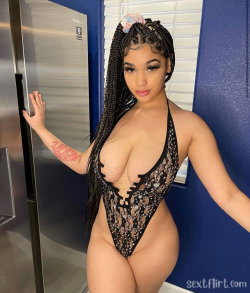 A curvy woman with long braided hair poses in a black lace bodysuit in a kitchen, hand on a stainless steel