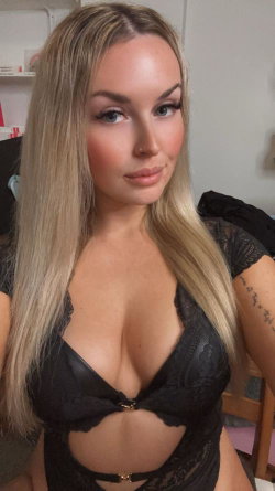 Blonde woman with long hair wearing black lace lingerie, posing indoors with visible arm tattoo.