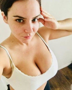 Brunette woman in white lace-trimmed bralette, posing closely with hand near head, indoors.