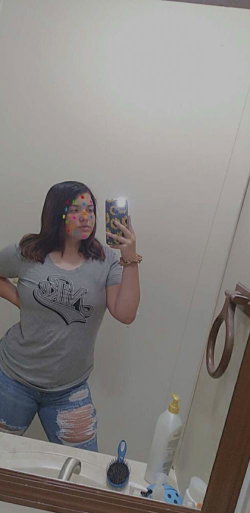 Woman with shoulder-length dark hair takes mirror selfie in gray t-shirt and ripped jeans, holding phone with colorful