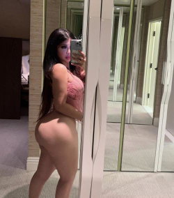 Curvy woman with long dark hair takes mirror selfie in pink lace top, bare bottom visible, standing in hotel room