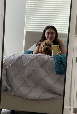 Brunette woman in a tank top takes a mirror selfie on a bed with a small dog, holding a phone.
