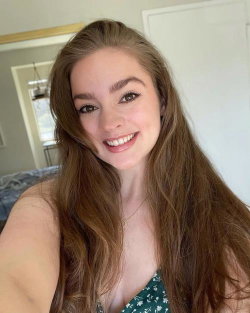 A smiling woman with long brown hair takes a selfie indoors, wearing a green floral top, with a bed and doorway visible