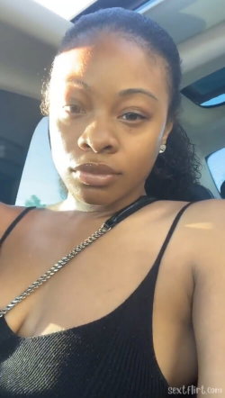 A Black woman with curly hair pulled back, wearing a black tank top and a chain strap, takes a selfie inside a car.