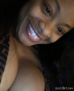 A smiling woman with dark skin and long hair, wearing a patterned top, captured in a close-up selfie with bare