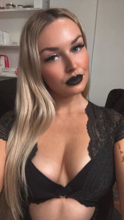 Blonde woman with black lipstick and lace top, posing indoors with visible tattoo on arm.