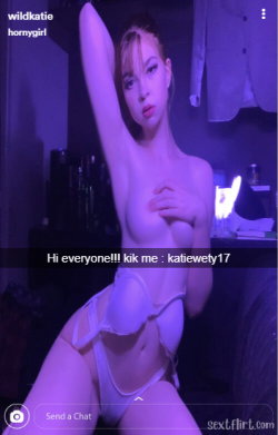 A woman with light brown hair poses topless in a dimly lit room, wearing white lingerie and holding one arm raised