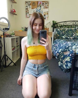 A young woman with pastel green-tipped hair takes a mirror selfie in a yellow crop top and denim shorts, kneeling on