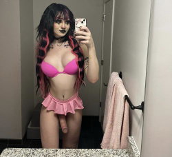 Person with black and pink hair in a pink bikini top and ruffled skirt, holding phone for mirror selfie in bathroom.