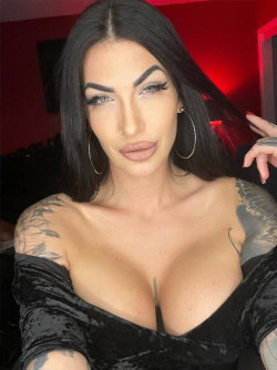 Brunette woman with tattoos on shoulders and arms, wearing a black off-the-shoulder top, posing in a red-lit room.