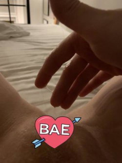 Close-up of a person's feet on a bed with a hand reaching toward them, featuring a pink heart sticker with 'BAE' text.