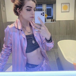 A woman with styled hair takes a mirror selfie in a bathroom, wearing a pink satin robe over a crop top and lace