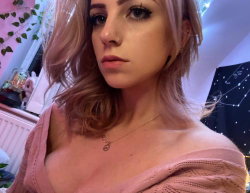A woman with pastel pink hair and a nose ring wears a loose pink sweater off one shoulder, posing indoors near plants
