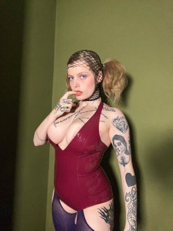 A woman with blonde hair in a ponytail, wearing a red latex top and purple thigh-highs, poses against a green wall with