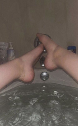 Close-up of bare feet resting on a bathtub edge, with water rippling below and bathroom fixtures in the background.