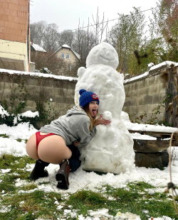 A woman in a gray hoodie and red thong crouches beside a snowman, holding a snowball, in a snowy backyard with a stone