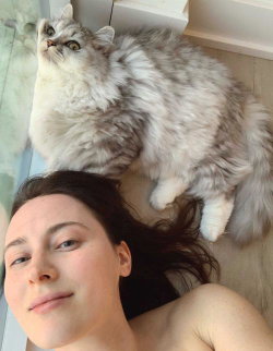 A woman with dark hair lies on her back, partially bare-chested, next to a fluffy gray cat on a light-colored floor.