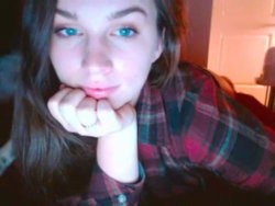 A young woman with long dark hair and blue eyes rests her chin on her hands, wearing a red and black plaid shirt