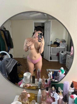 A curvy woman in pink thong underwear takes a selfie in a cluttered bedroom.