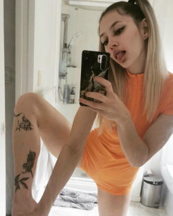 Blonde woman in orange dress takes mirror selfie in bathroom, showing leg tattoos and playful expression.