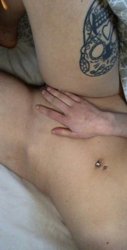 Close-up of a person's torso and thigh with a detailed skull tattoo, navel piercing, and hand resting on skin, lying on