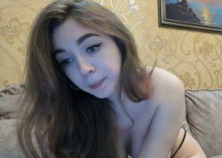 Brunette woman with long hair, topless, leaning on a couch in a room with patterned wallpaper and a framed picture.