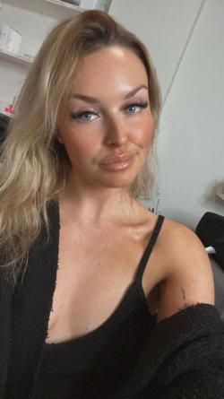 Blonde woman with blue eyes in a black top and cardigan, taking a selfie indoors with soft lighting.