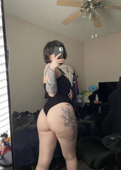 Curvy woman with tattoos takes mirror selfie in black bodysuit, showing back and hip tattoo in bedroom setting.