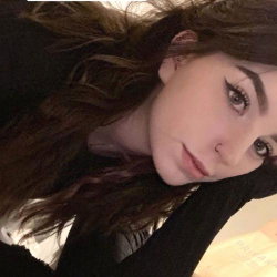 Brunette woman with wavy hair and dark eyeliner rests her head on a black sleeve, looking at the camera in a close-up