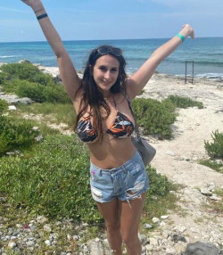A woman with long dark hair in a patterned bikini top and ripped denim shorts stands on a rocky beach with arms raised,