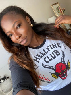A woman with long dark hair and a graphic tee takes a selfie indoors, holding her hair while looking at the camera.