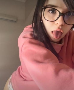A young woman with dark hair and glasses sticks her tongue out while wearing a pink hoodie, taking a close-up selfie