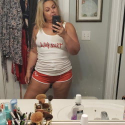 Blonde woman in white tank top and red shorts takes mirror selfie in bathroom, showing tattoos on arm and side.
