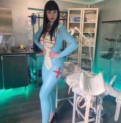 A woman in a tight blue latex nurse outfit with red crosses poses confidently in a medical-themed room with equipment