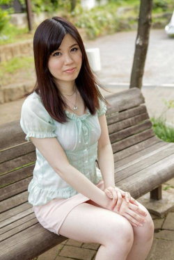 A woman with shoulder-length dark hair sits on a wooden bench outdoors, wearing a light green ruffled top and pink