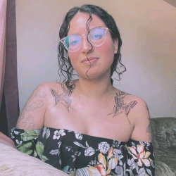 A woman with dark curly hair and glasses wears an off-shoulder floral top, showing tattoos on her shoulders while
