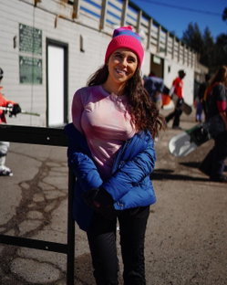 A smiling woman with long dark hair wears a pink beanie, blue puffer jacket, and tight pink top while leaning on a
