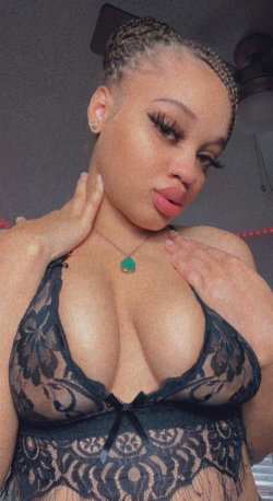 A woman with braided hair and bold makeup wears a black lace lingerie top, posing indoors with hands on her neck and