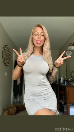 Blonde woman in a tight gray dress making peace signs and sticking out her tongue, standing in a bathroom with visible