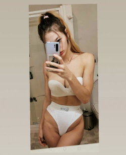 A woman with long hair in a high ponytail takes a mirror selfie in white lingerie, showing a curvy figure in a bathroom
