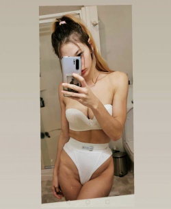 A woman with long hair in a high ponytail takes a mirror selfie in white lingerie, standing in a bathroom.