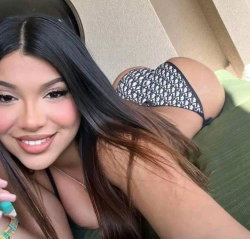 Brunette woman with long hair smiles in a selfie, wearing patterned thong underwear while lying on a green surface.