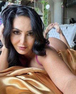 Brunette woman with wavy hair lying on a gold satin sheet, wearing a pink top, in a decorated indoor setting.