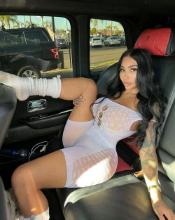 A woman with long dark hair and tattoos sits in a car, wearing a sheer white dress and white socks, posing with one leg