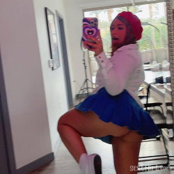 A woman in a white shirt and blue skirt takes a mirror selfie, wearing a red beret and white socks, indoors near a