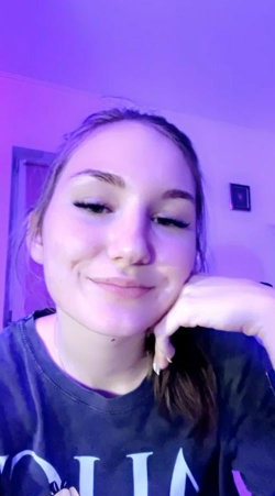 A young woman with brown hair and makeup smiles softly while resting her chin on her hand in a room with purple
