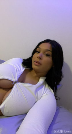 A curvy woman with dark hair lies on a bed, wearing a white zip-up top that exposes her bare chest.