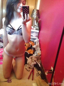 A young woman with long dark hair kneels on the floor in a lace bra and light-colored underwear, taking a mirror selfie