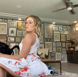 A blonde woman in a floral dress sits sideways on a couch, wearing glasses, in a room with framed artwork on the walls.